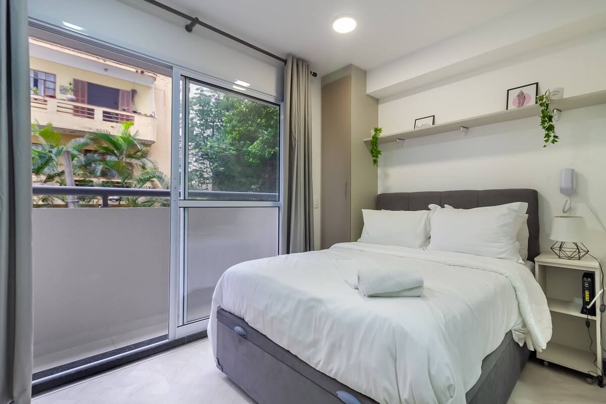 Ein modernes Schlafzimmer mit einer großen Glasschiebetür, die auf einen Balkon führt, einem Bett mit weißen Bettwäsche, einem Nachttisch mit Lampe und einem Regal mit Dekoration.
