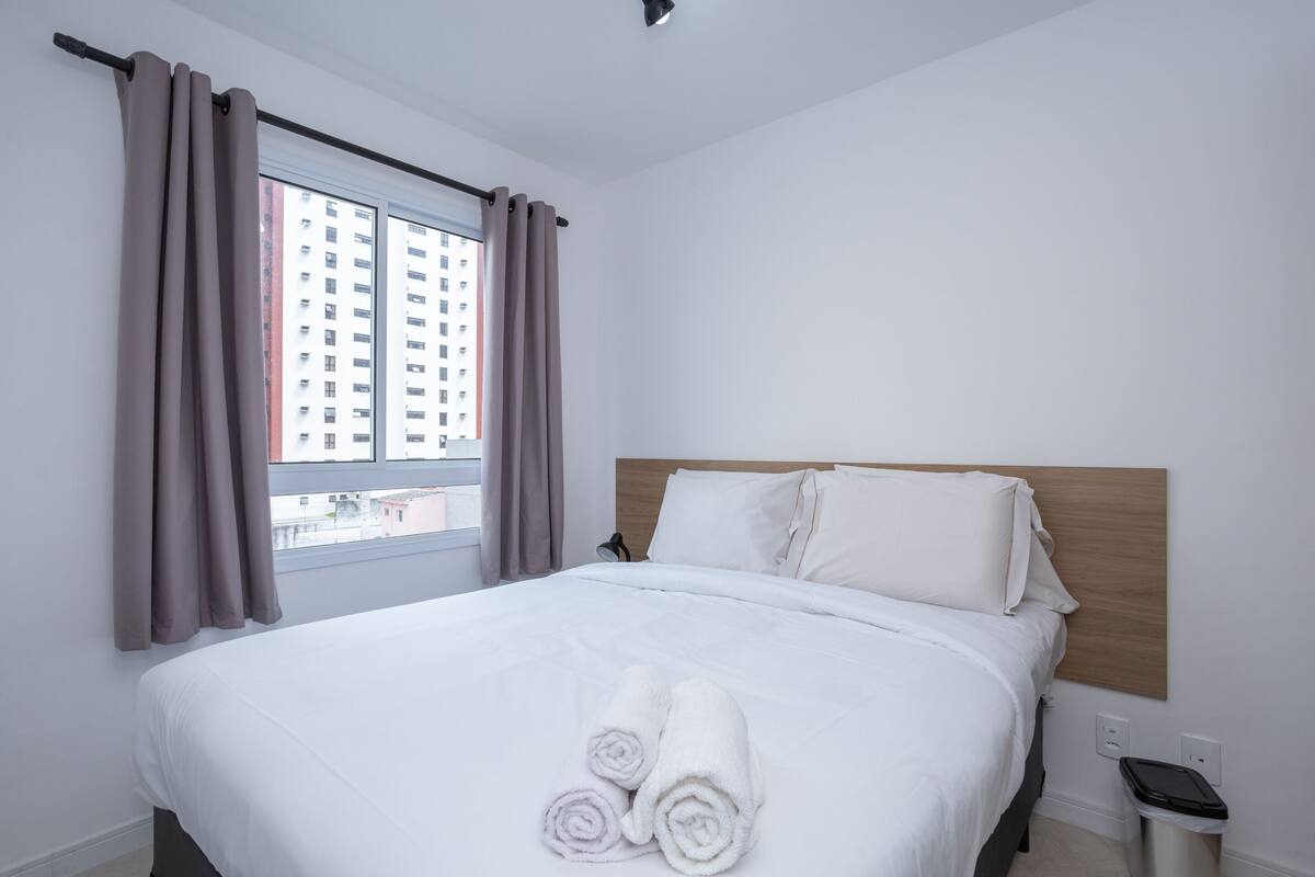 Ein Hotelzimmer mit Bett, einem Vorhangfenster, einem Nachttisch und Blick auf Gebäude.