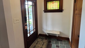 Un passadís amb una porta de fusta, una petita finestra amb vitralls, una banca blanca i una catifes amb motius.