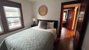 Un dormitori amb un llit gran, una làmpada de nit, un penjoll rodó a la paret i una vista a través d'una porta oberta cap a una altra habitació.