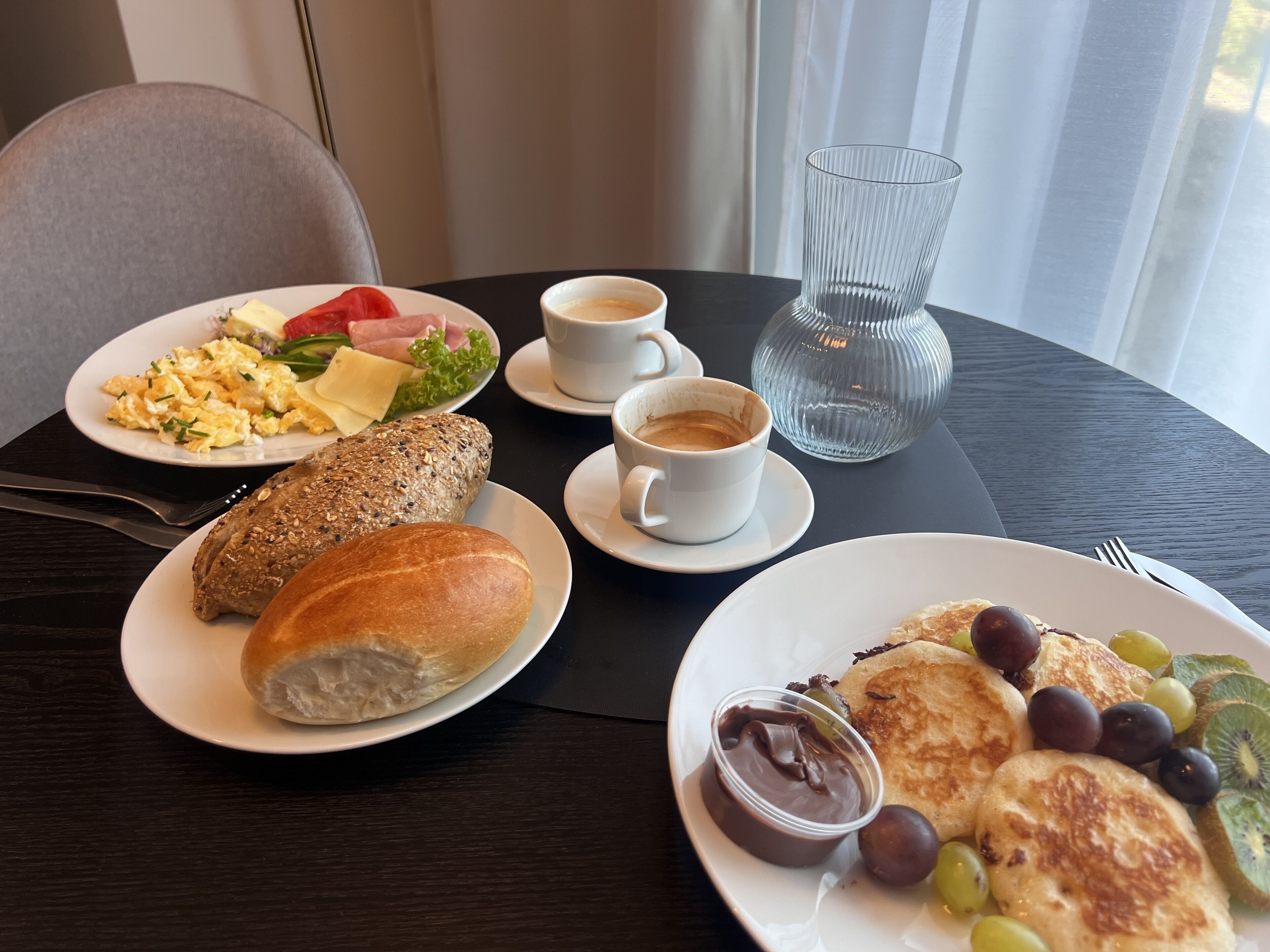 A breakfast table with plates of scrambled eggs, ham, and toast, two cups of coffee, and a glass vase.