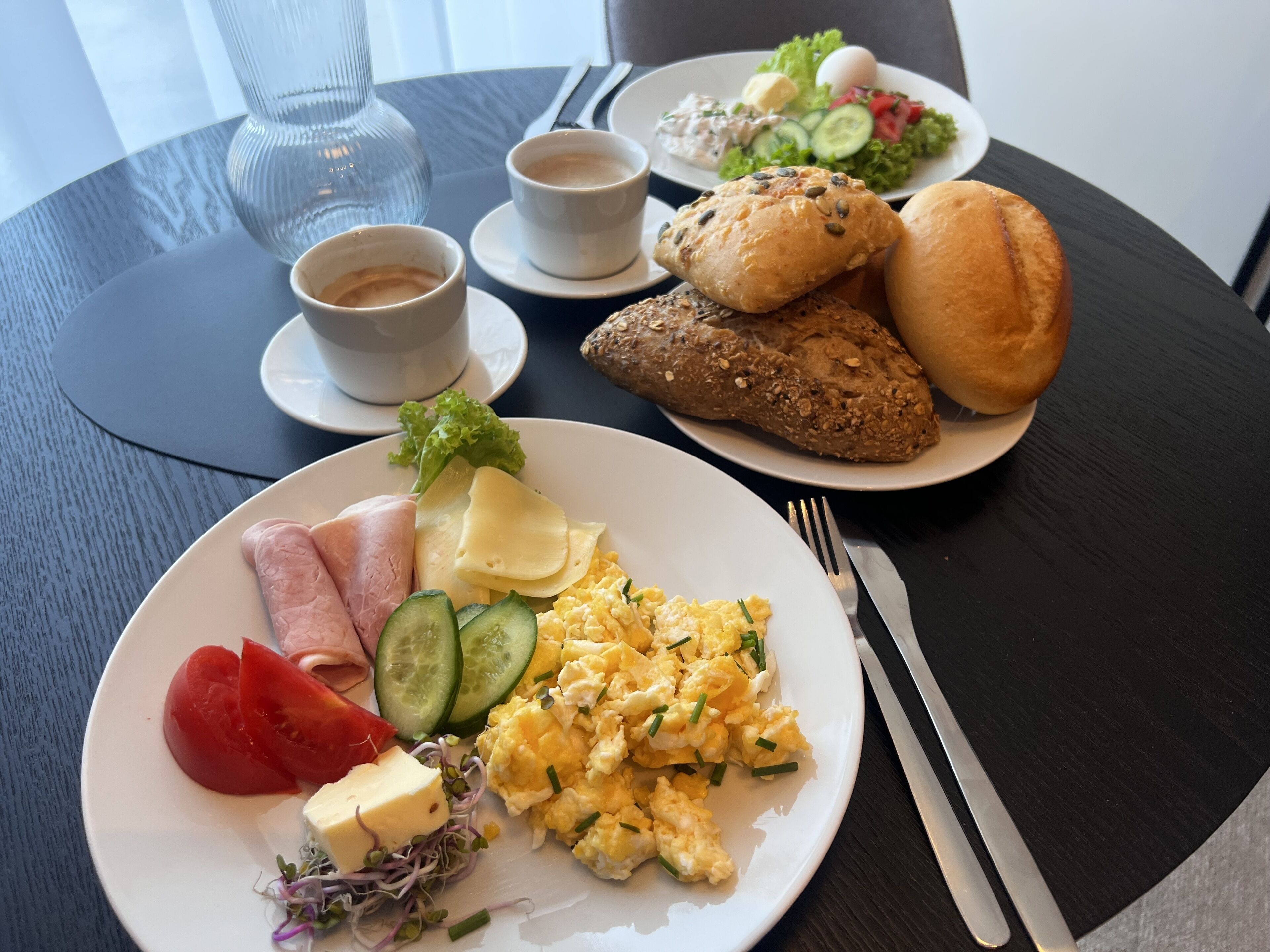 A breakfast setting with plates of eggs, ham, cheese, and vegetables, along with bread and coffee cups on a table.