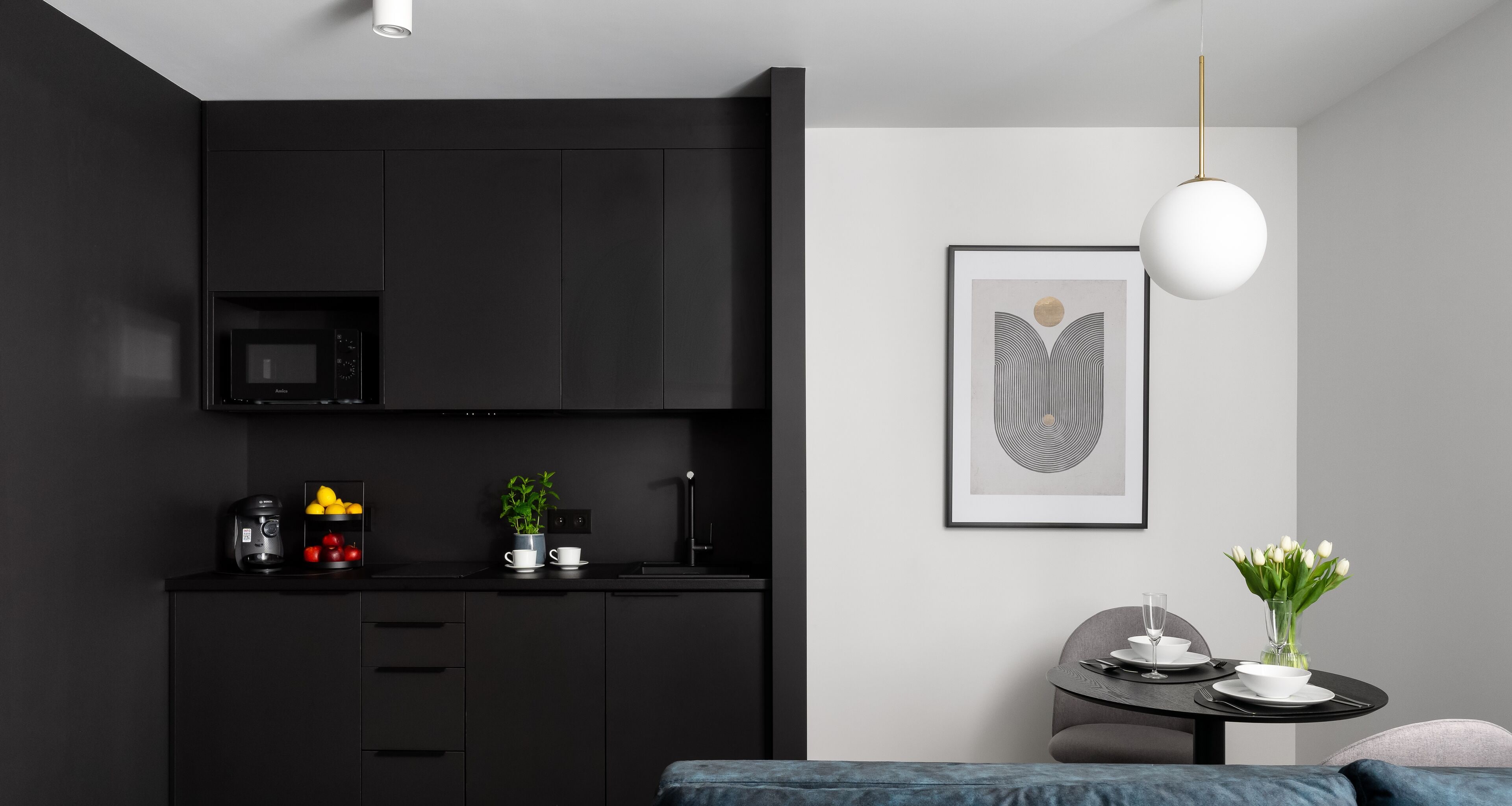 A modern kitchen with black cabinets, a microwave, and a coffee machine. There is a dining area with a table, chairs, and a vase of flowers.
