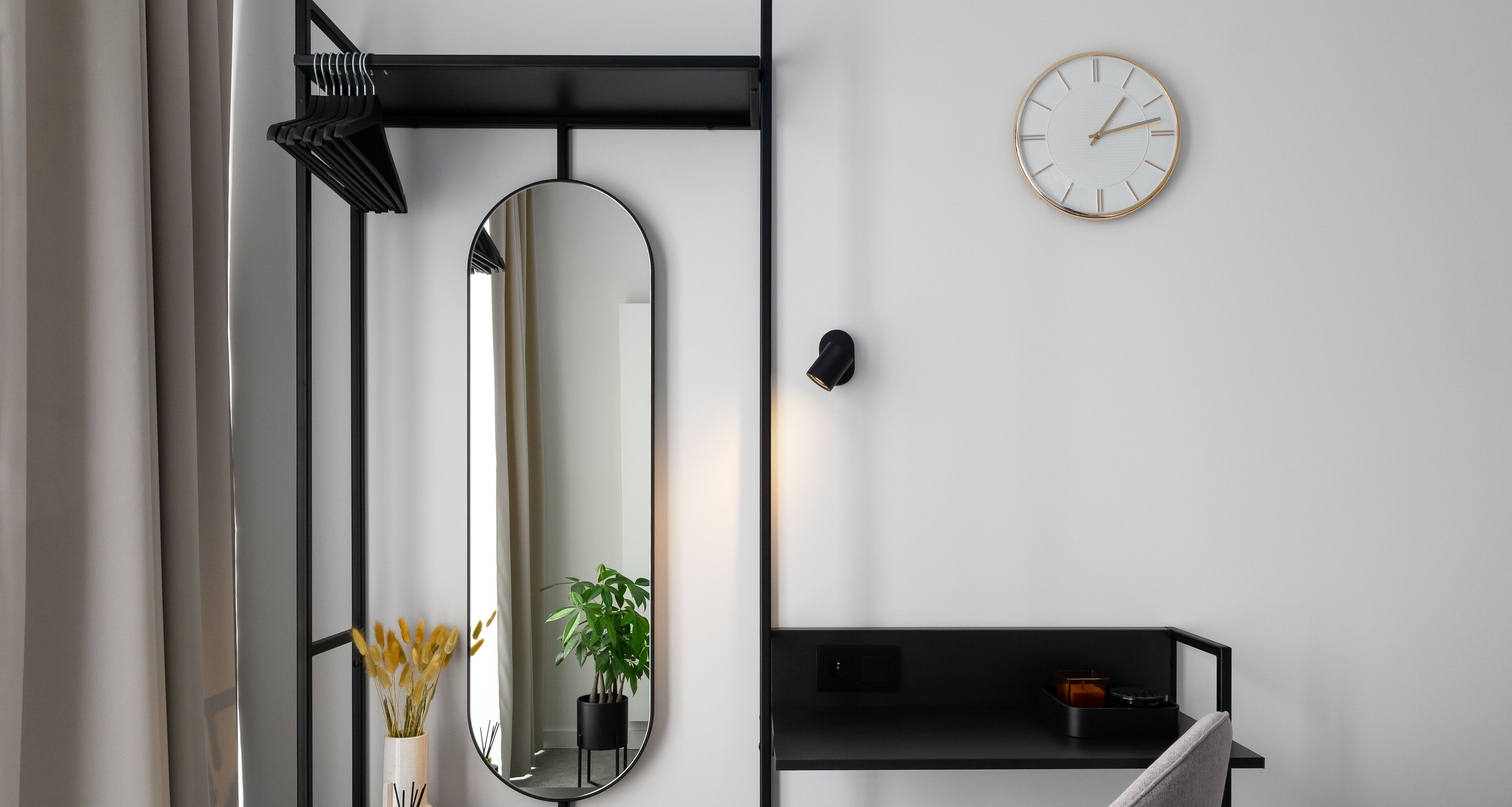 A modern bathroom with a large oval mirror, a round wall clock, a potted plant, and a black shelf with a small orange container.