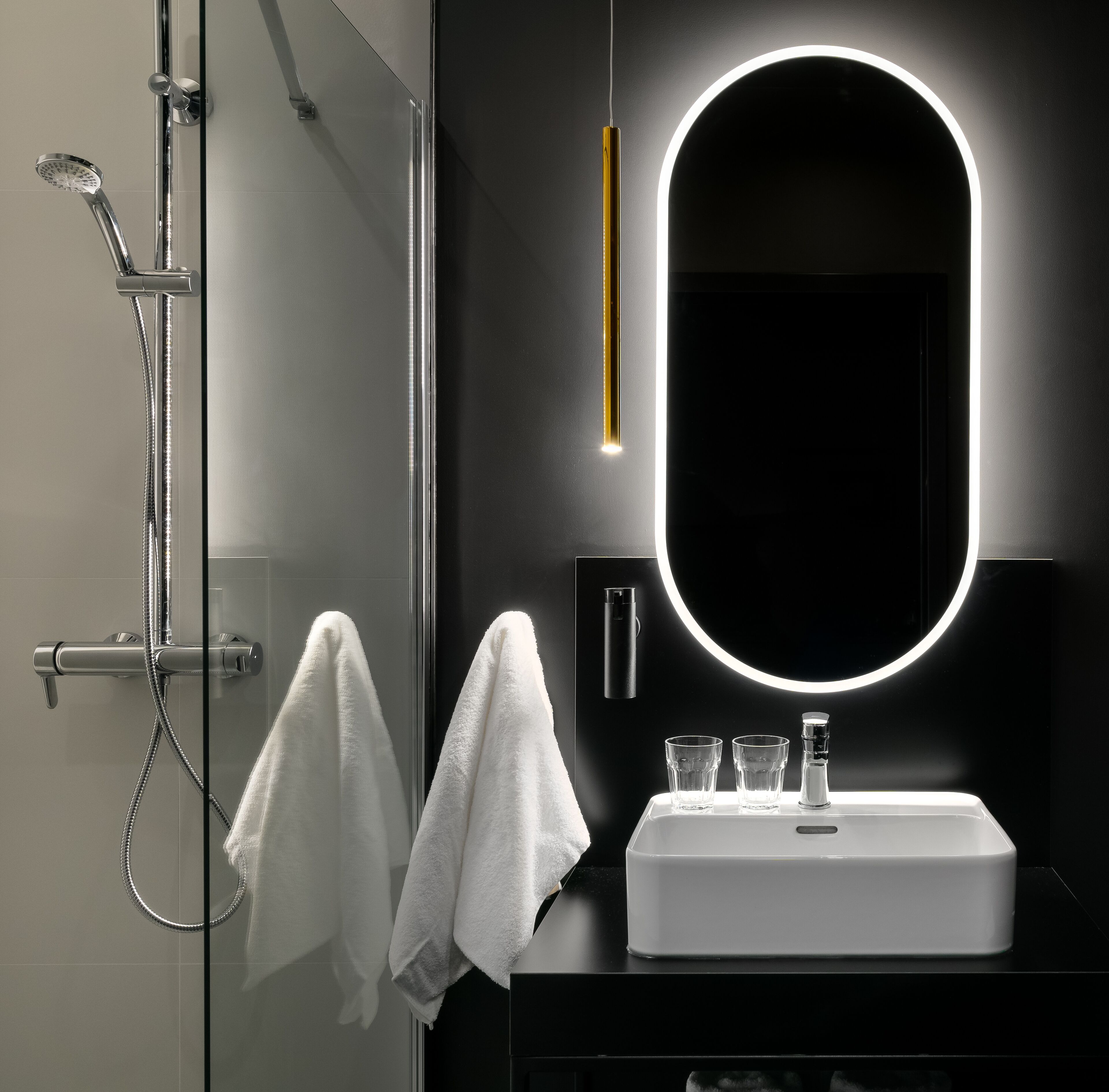 Un baño moderno con cabina de ducha acristalada, un lavamanos blanco y un espejo grande con luz circular.