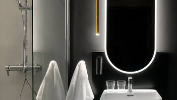 Un baño moderno con cabina de ducha acristalada, un lavamanos blanco y un espejo grande con luz circular.