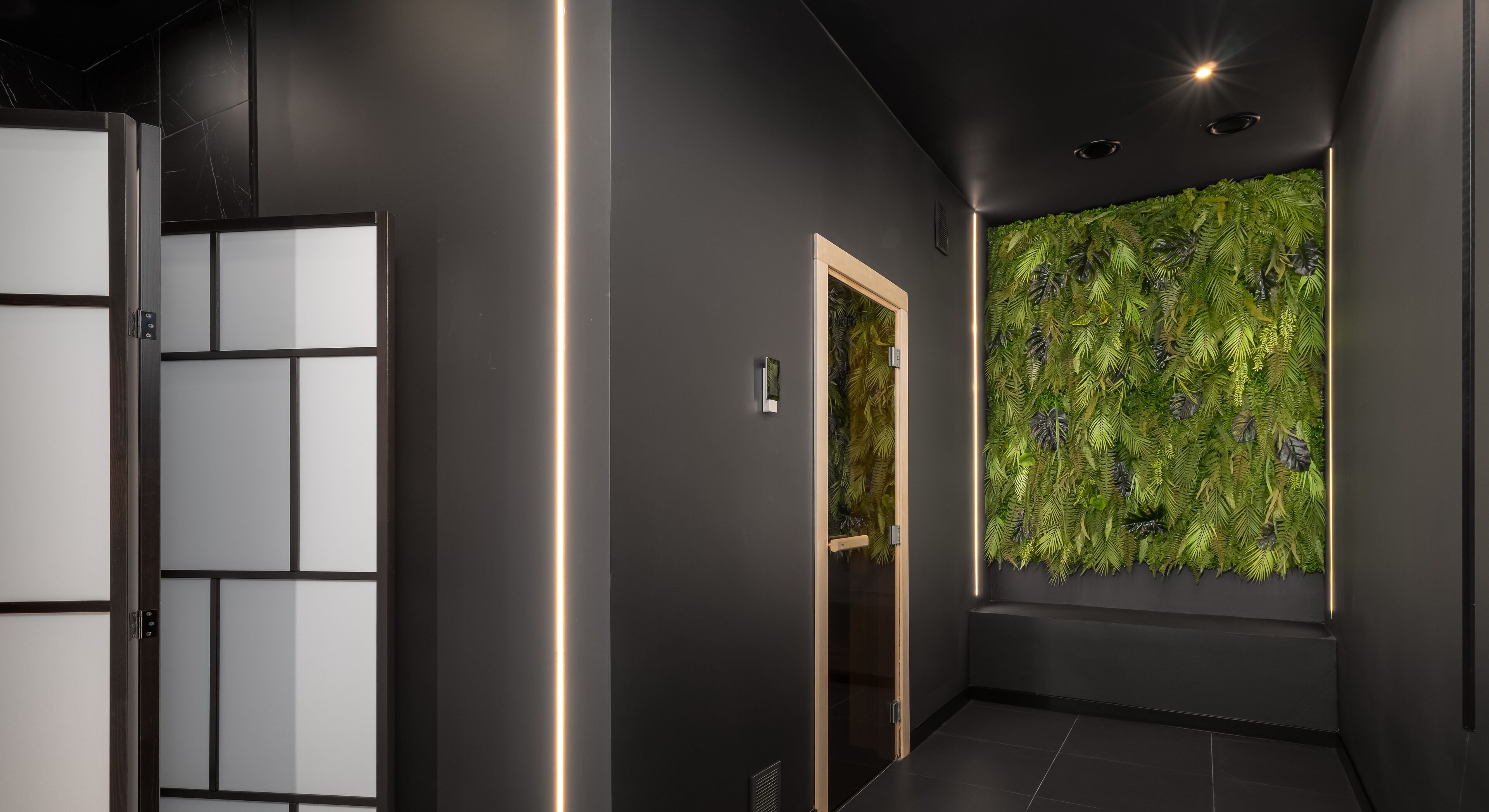 A modern hallway with a dark wall, a glass door, and a green leafy wall.
