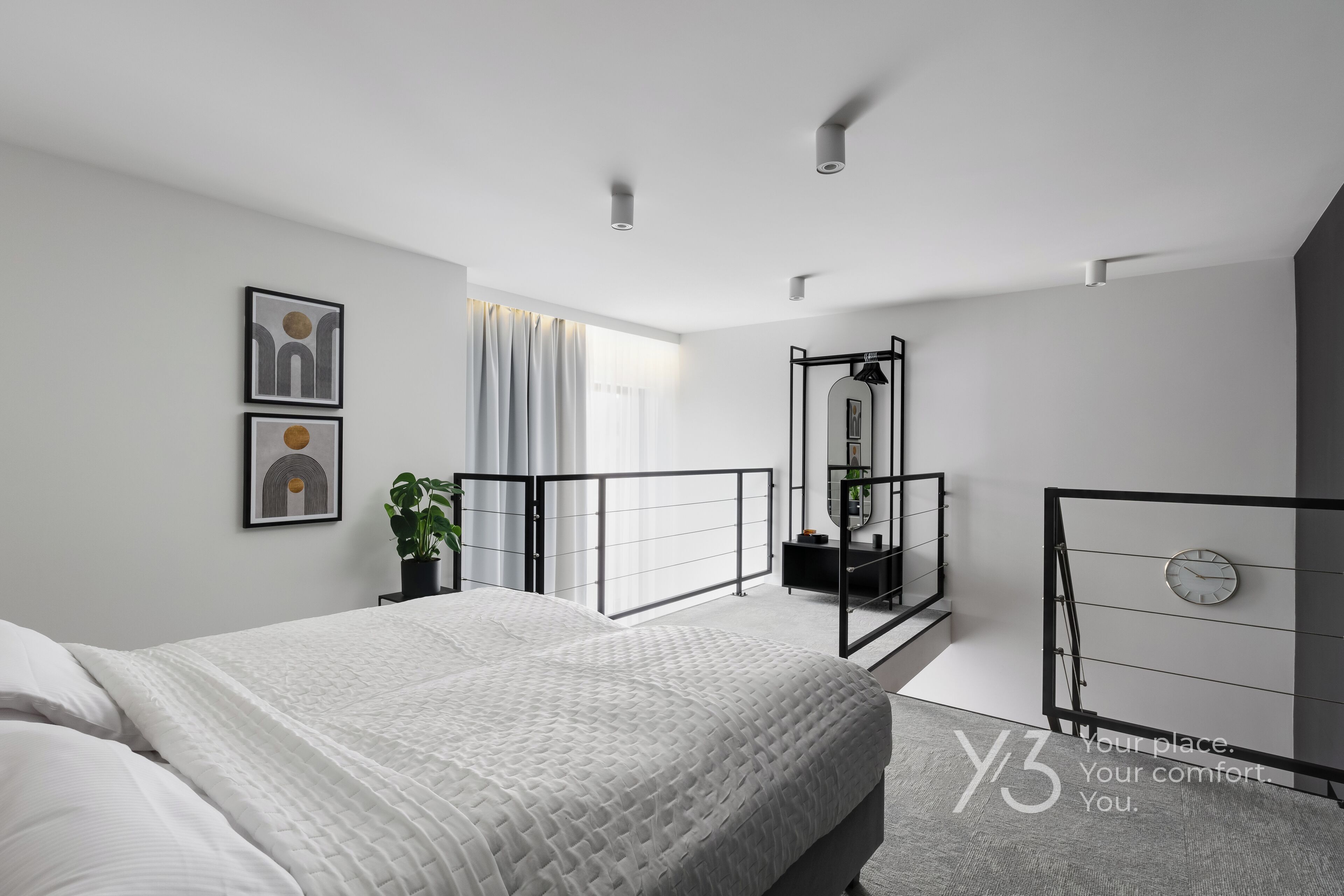 A modern bedroom with a large bed, a bedside table with a plant, a mirror, and a clock on the wall.