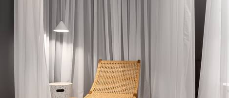 Una silla de mimbre para relajarse en una habitación con cortinas blancas translúcidas y un taburete pequeño y redondo.