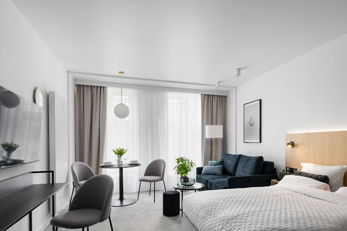 A modern hotel room with a large bed, a desk with a chair, a small table with a chair, a sofa, and a TV mounted on the wall.