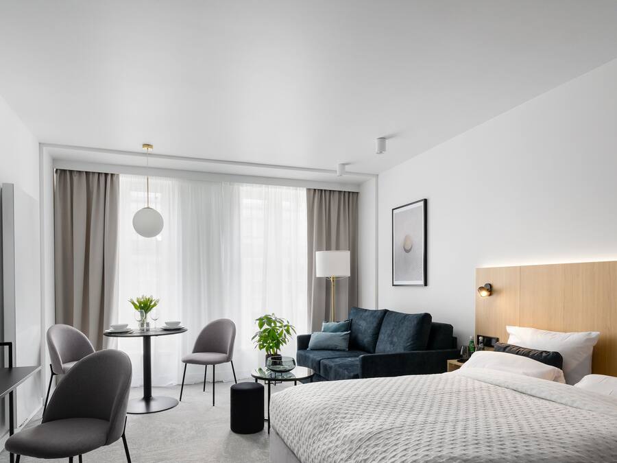 A modern hotel room with a large bed, a desk with a chair, a small table with a chair, a sofa, and a TV mounted on the wall.