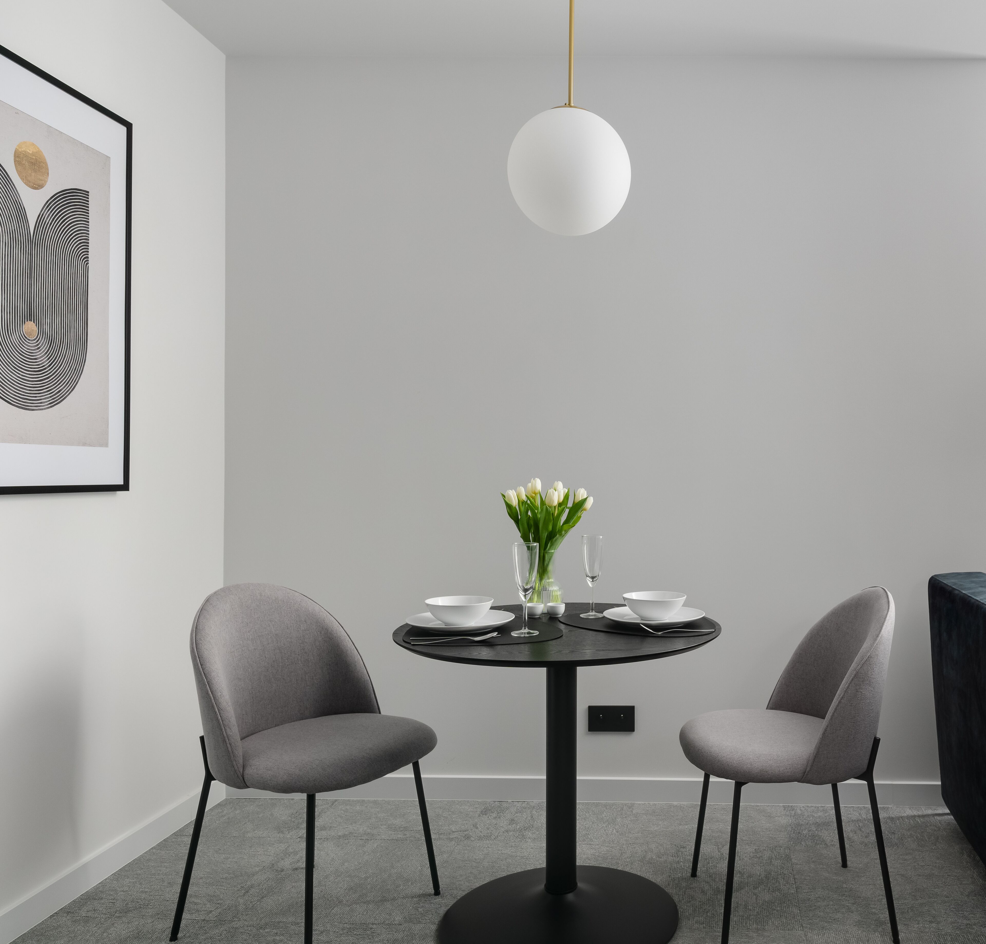 Un espace repas moderne avec une table ronde, deux chaises grises, un luminaire suspendu blanc et une œuvre d’art abstraite encadrée au mur.