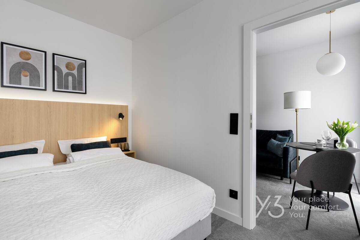 A modern hotel room with a large bed, a wooden headboard, a desk with a chair, and a dining table with a vase of flowers.