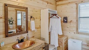 Ein Badezimmer einer Holzhütte mit einem Holzwaschbecken, einem Spiegel, einem Handtuchhalter und einem weißen Bademantel, der an einem Gestell hängt.