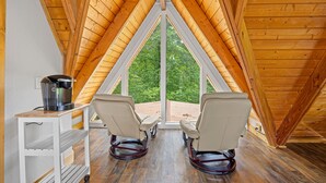 A room with a wooden sloped ceiling, two reclining chairs, a small table with a kettle, and a large window offering a view of greenery. - Selah (Gatlinburg)