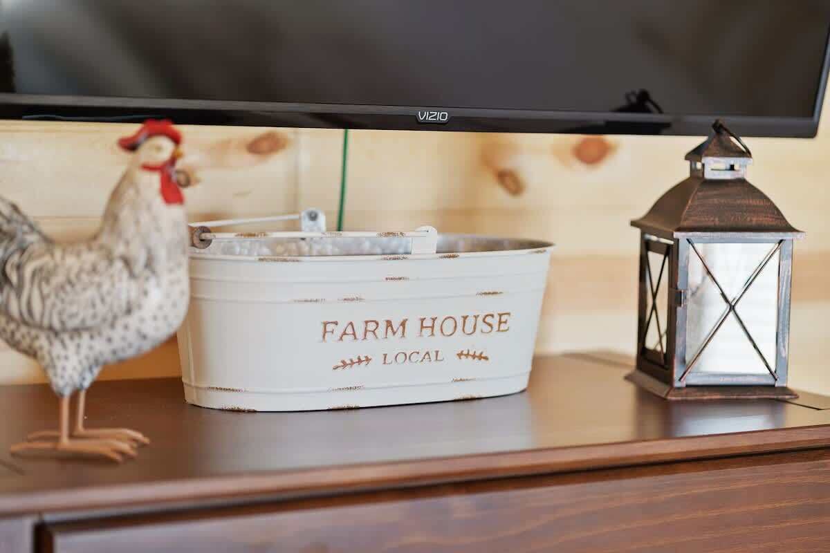 Un téléviseur, un conteneur blanc portant l’inscription « PRODUITS DE LA FERME », une lanterne en métal et un poulet posé sur une surface en bois.