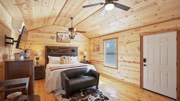 A cozy wooden cabin room with a bed, a leather sofa, a dining table, and a ceiling fan. - Gorgeous Cabin on Main-steps to Pacific War Museum (Fredericksburg)