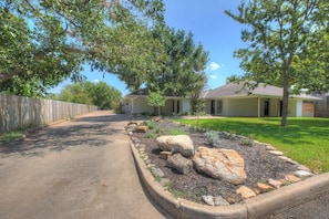 A residential driveway leading to a house with a covered porch, a stone garden bed, and a wooden fence. - Wanderlust #1 Hot Tub Shared Outdoor Space (Fredericksburg)