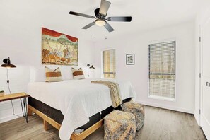 A bedroom with a bed, a ceiling fan, a window with blinds, a bedside table, and a wall painting. - Wanderlust #1 Hot Tub Shared Outdoor Space (Fredericksburg)