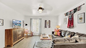 A modern living room with a leather sofa, a wooden cabinet, and a dining table. There is a door with curtains and a window with blinds. - Wanderlust #1 Hot Tub Shared Outdoor Space (Fredericksburg)