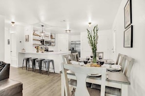 Uma cozinha moderna com armários brancos, uma ilha central com banquetas e uma área de refeições com mesa e cadeiras de vidro.
