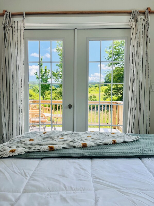 Une chambre avec un grand lit, une vue sur un paysage verdoyant grâce à de grandes portes-fenêtres, et une terrasse en bois à l’extérieur.