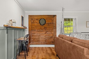 Une pièce avec un mur en bois sur lequel est accroché un jeu de fléchettes, un canapé, une fenêtre avec des rideaux et une table avec des chaises.