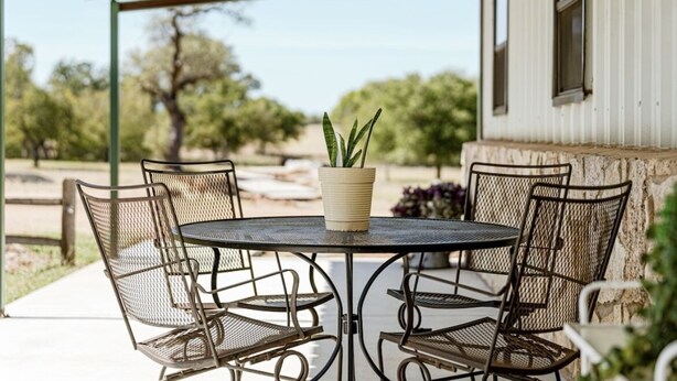 A covered patio with a round table and four chairs, a potted plant on the table, and a view of trees and a building in the background. - Faulkner Ranch Bunkhouse (Llano)