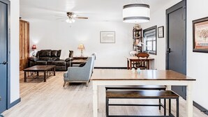 A living room with a sofa, armchair, coffee table, and a dining table with benches. There is a ceiling fan and a chandelier. - Faulkner Ranch Bunkhouse (Llano)