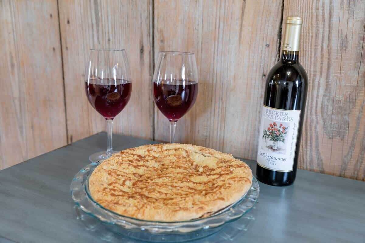 A glass pie on a clear plate, two glasses of red wine, and a bottle of wine on a wooden table.