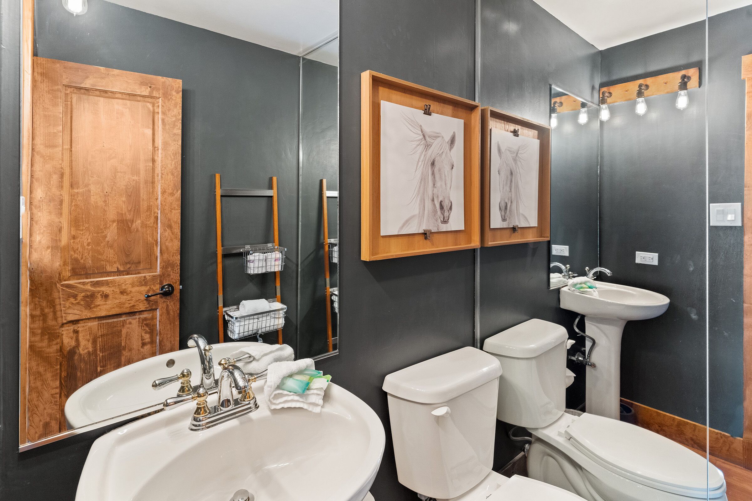 A bathroom with a large mirror, a wooden door, a white sink, two toilets, and framed artwork on the wall.