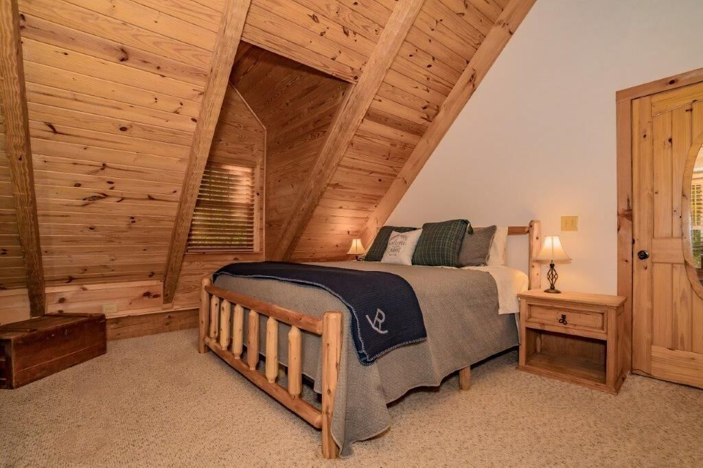 A bedroom with a sloped ceiling, wooden walls, a bed with a blue blanket, two bedside tables with lamps, and a wooden door.