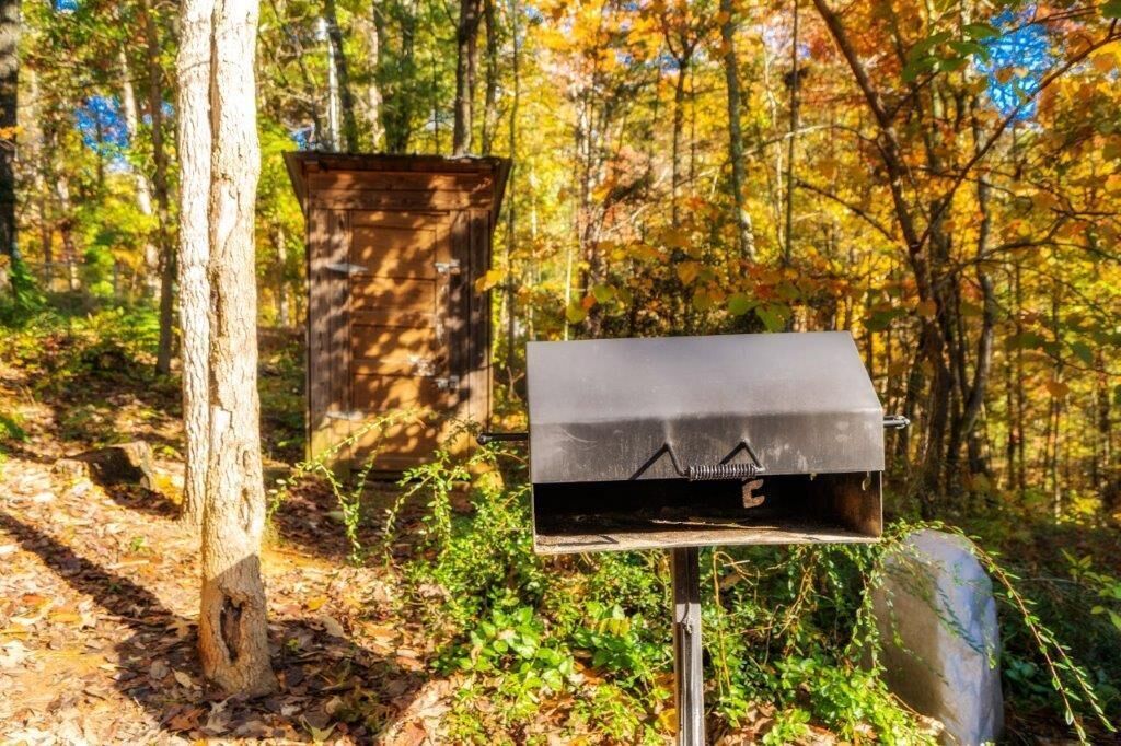 A metal grill box in a forested area with a wooden structure in the background.