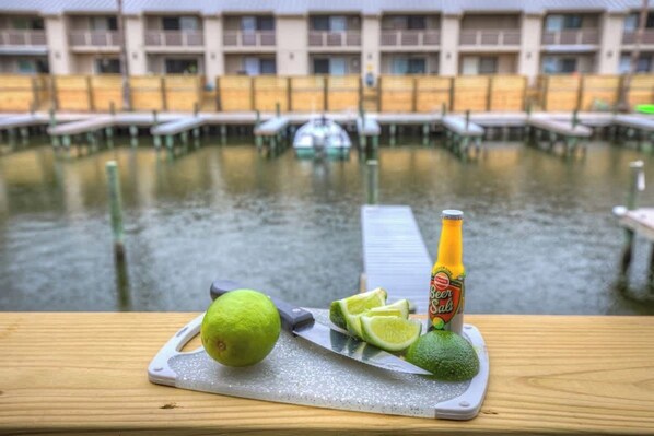 A table with a lime, a knife, and a bottle of beer on a dock by a calm body of water, with a building in the background.