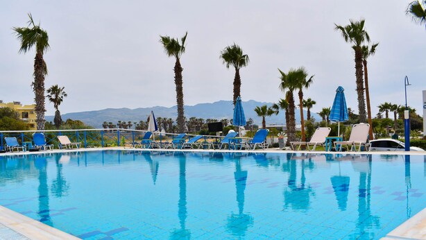 Ein Swimmingpool mit Liegestühlen und Sonnenschirmen, umgeben von Palmen und Bergen im Hintergrund.