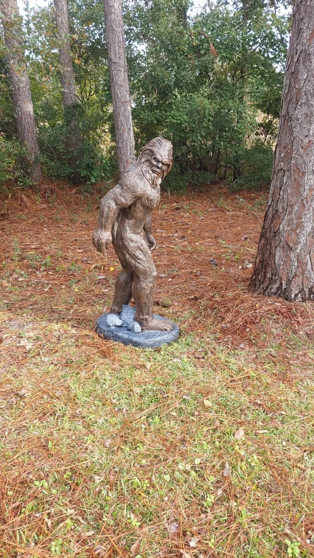 Una estatua de bronce de un oso en un área boscosa con agujas de pino en el suelo.