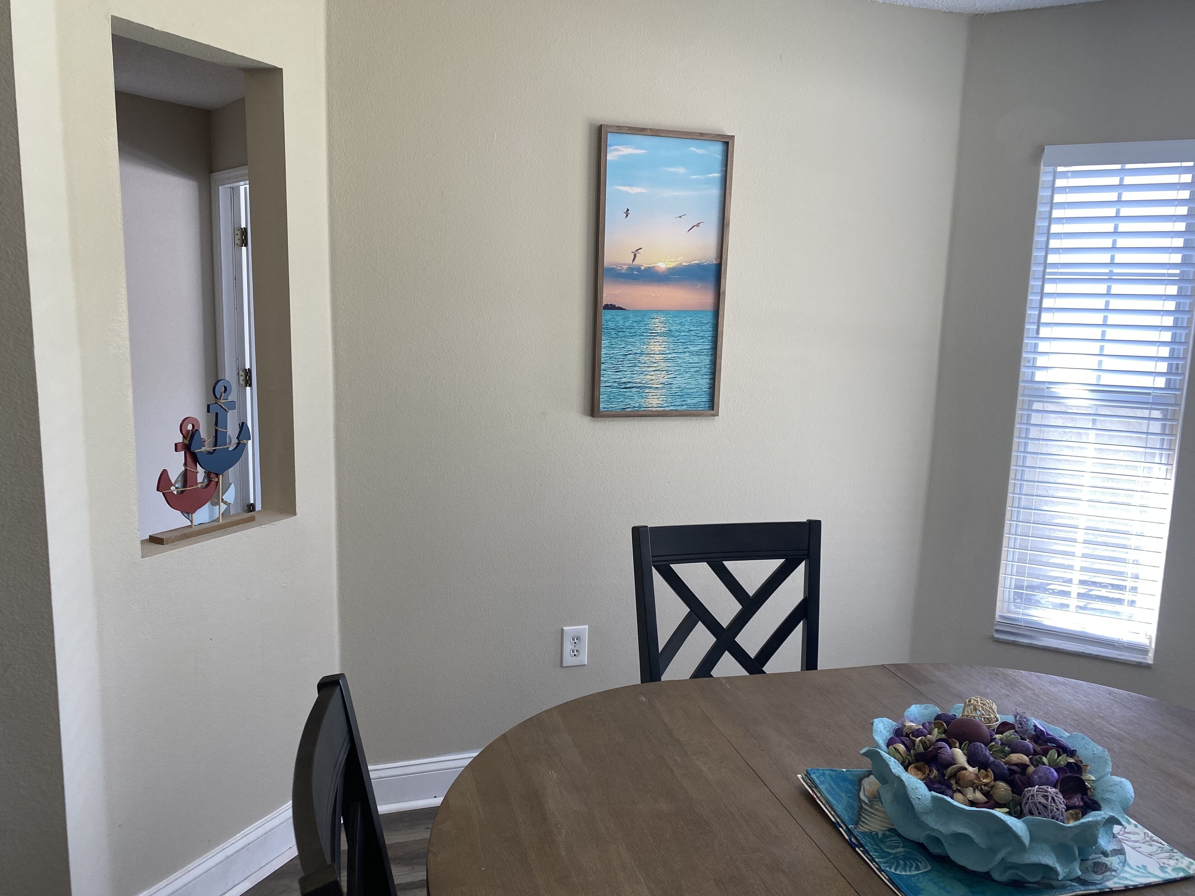 A dining area with a round table, a framed picture of a sunset over water, and a window with blinds.