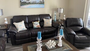 Een woonkamer met een bruine leren bank, decoratieve kussens, een bijzettafel met koraalsculpturen, een schilderij van een strandlandschap en een lamp op een nachtkastje.