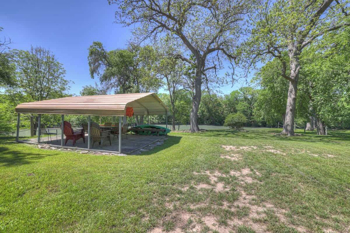A covered outdoor seating area with chairs and a table, surrounded by trees and grass.