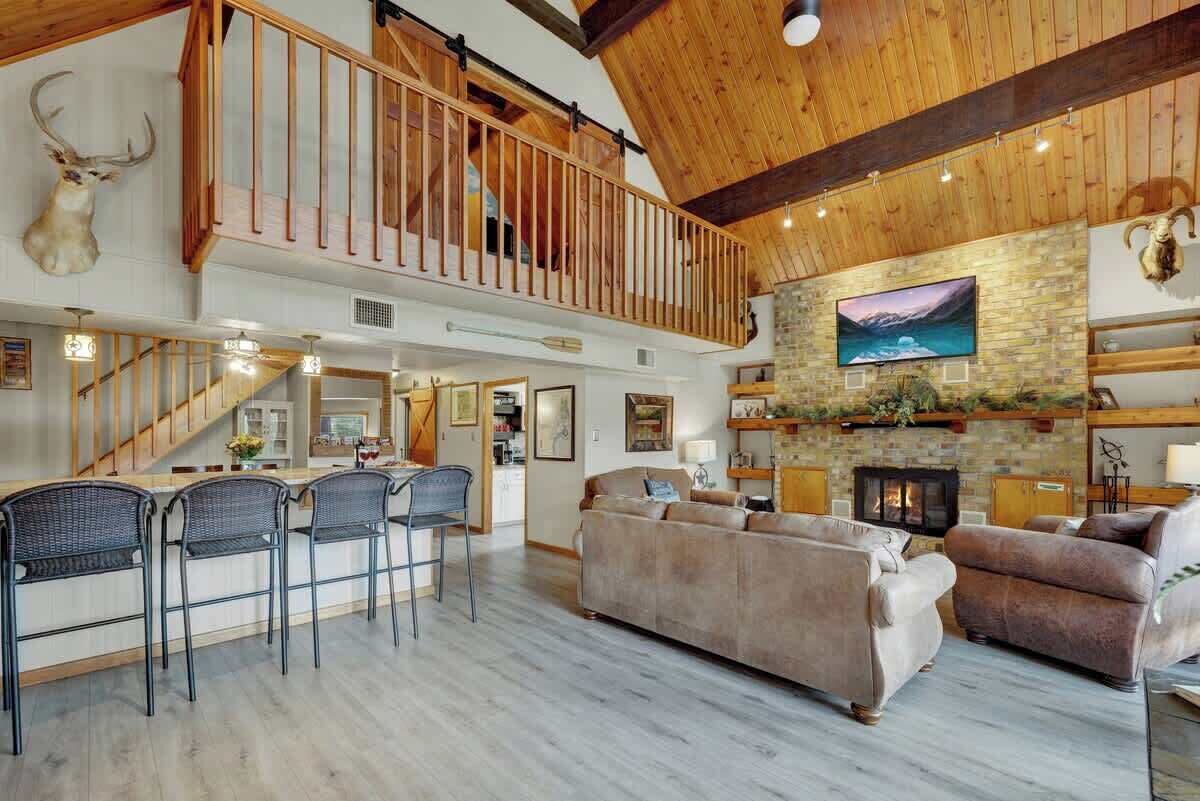 A spacious living area with a high ceiling, a fireplace, and a stone wall. There is a staircase leading to an upper level with a balcony.