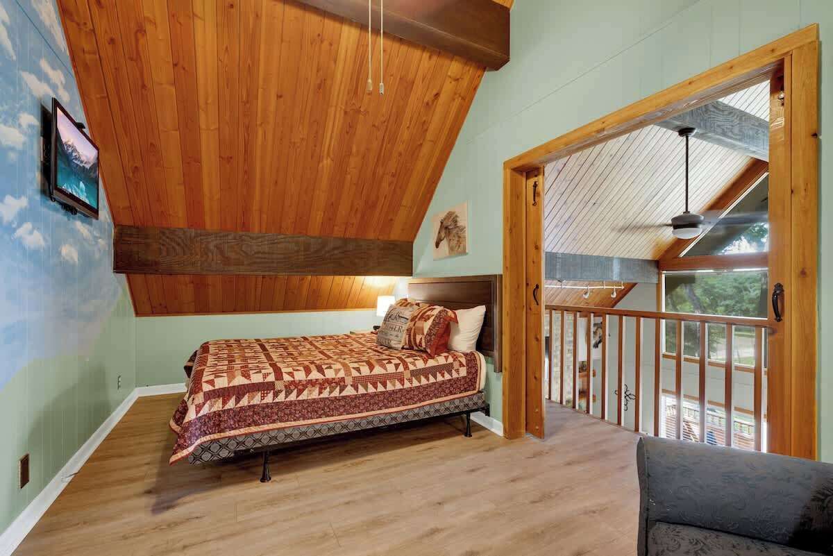 A bedroom with a wooden ceiling, a bed with patterned bedding, and a balcony with a railing.