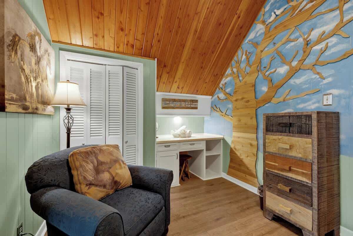 A cozy room with a wooden ceiling, a desk with a lamp, a chair, and a wall with a tree mural.