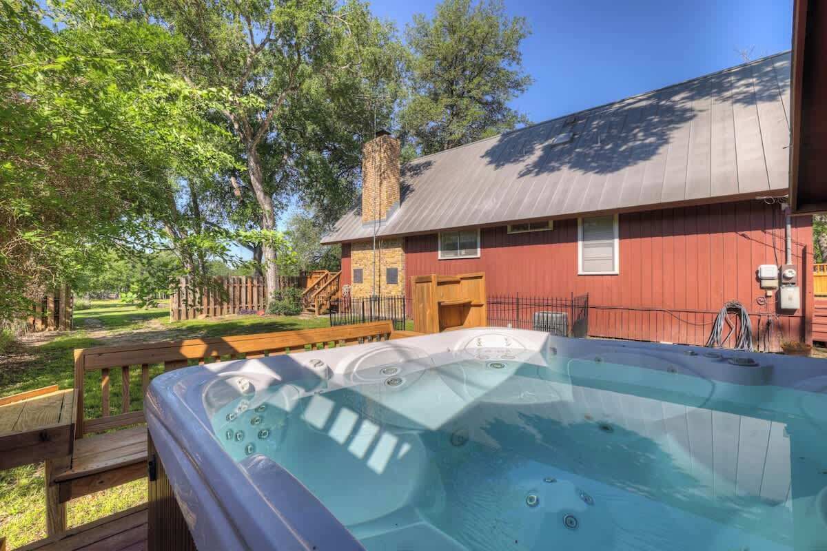 A hot tub in front of a red building with a chimney, surrounded by trees and a wooden deck.