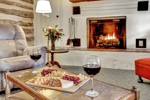 A cozy room with a fireplace, a sofa, a table with a cheese board and wine glasses. - Historic Log Cabin Retreat Near Town on 5 Acres! (Fredericksburg)