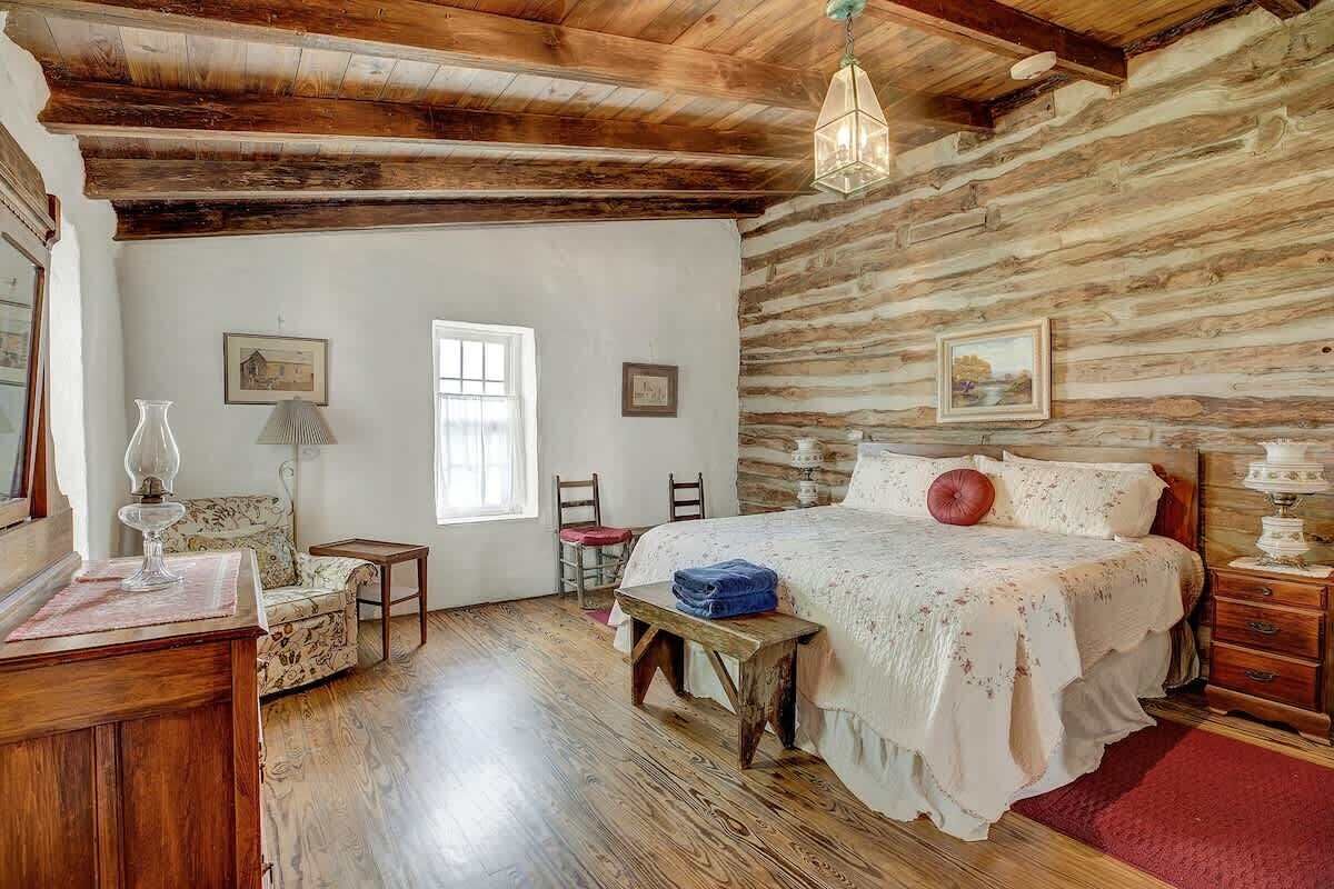 A rustic room with wooden walls, a bed with a floral bedspread, a wooden bench, a chair, a table, and a hanging light fixture.