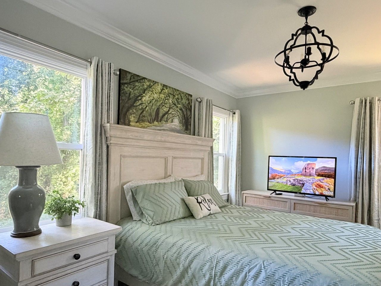 A bedroom with a bed, a nightstand, a lamp, a television, and a window with curtains.