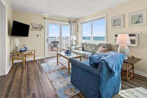 A living room with a view of the ocean, a television, and a balcony with outdoor furniture.
