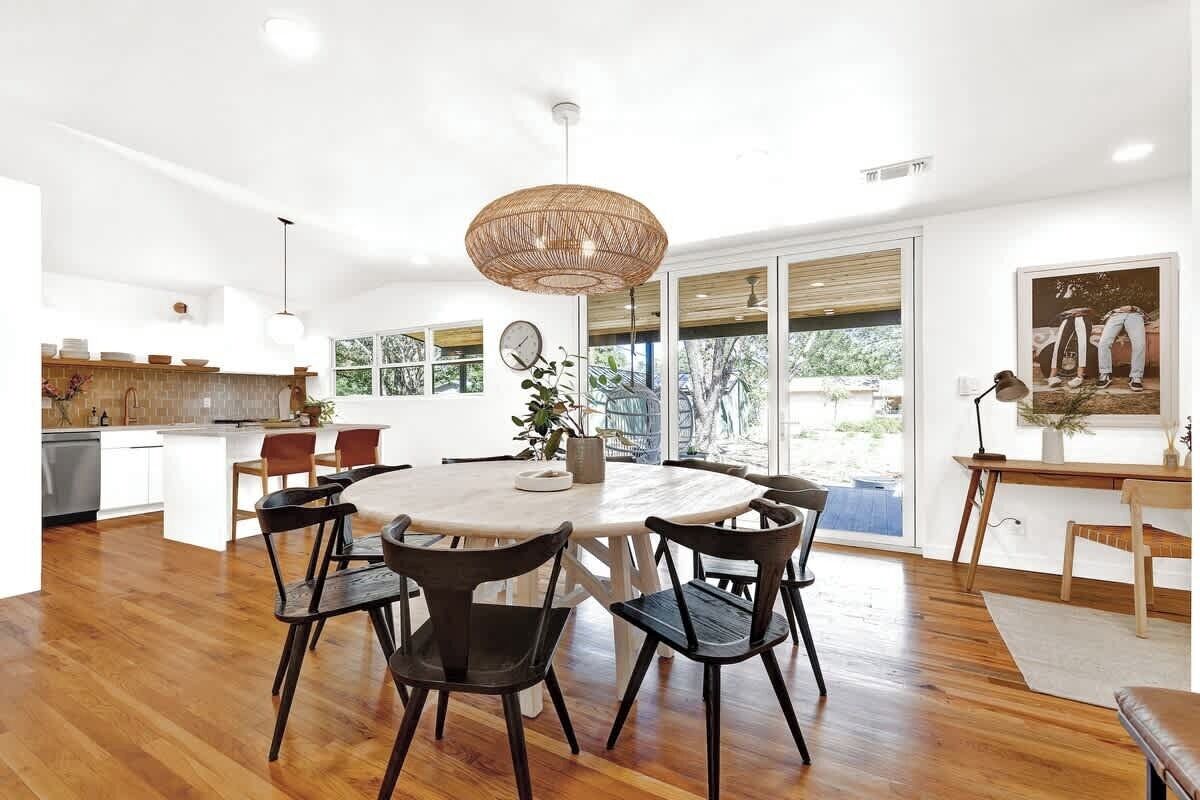 A spacious dining area with a round table and chairs, a large woven pendant light, and a view of the outdoors through large windows.
