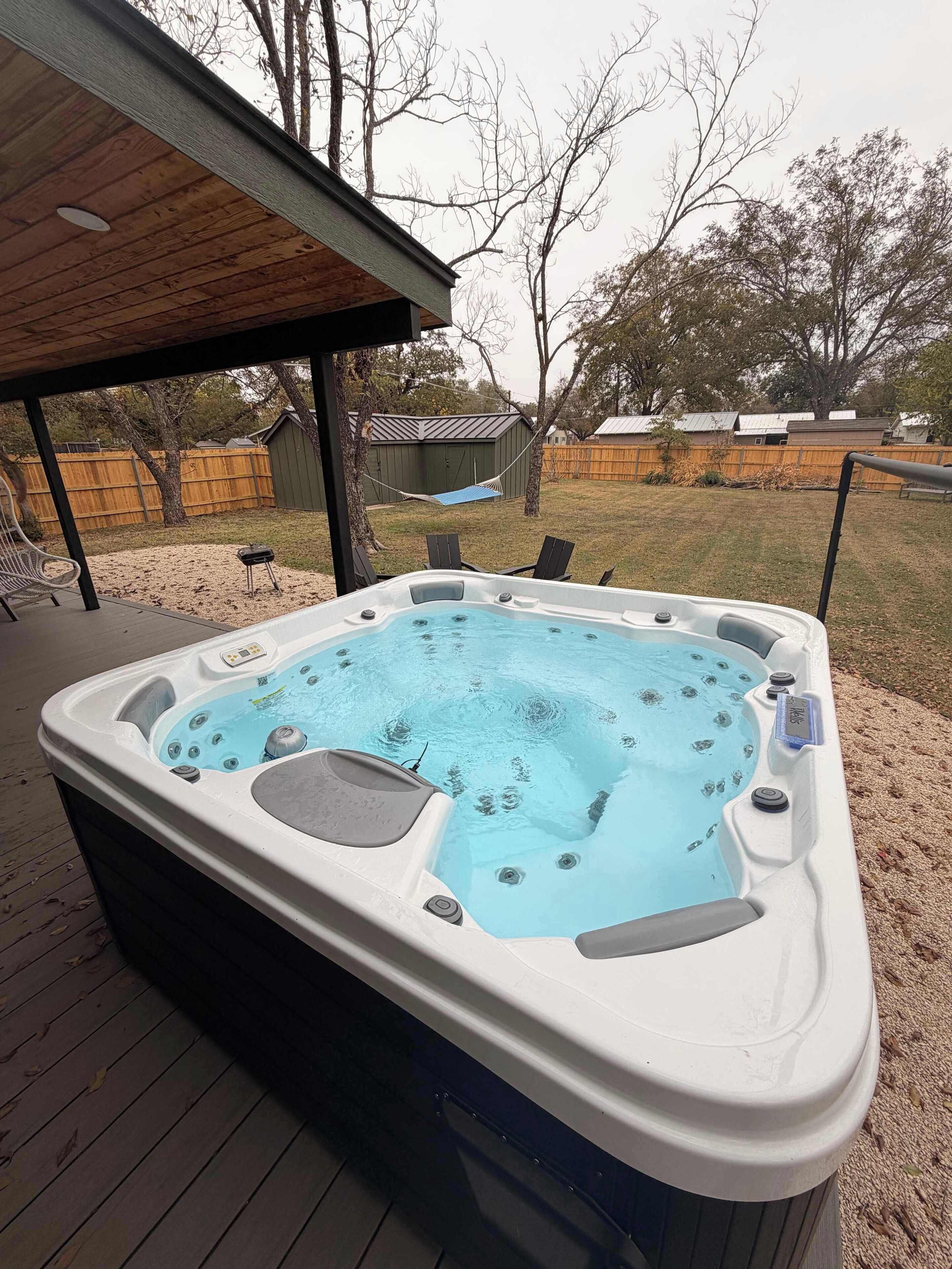Een hottub op een houten terras, met een overdekte ruimte erboven, omgeven door een omheind gazon en bomen.