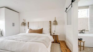 Ett modernt sovrum med en stor säng, sängbord, en hängande lampa och utsikt över ett badrum.